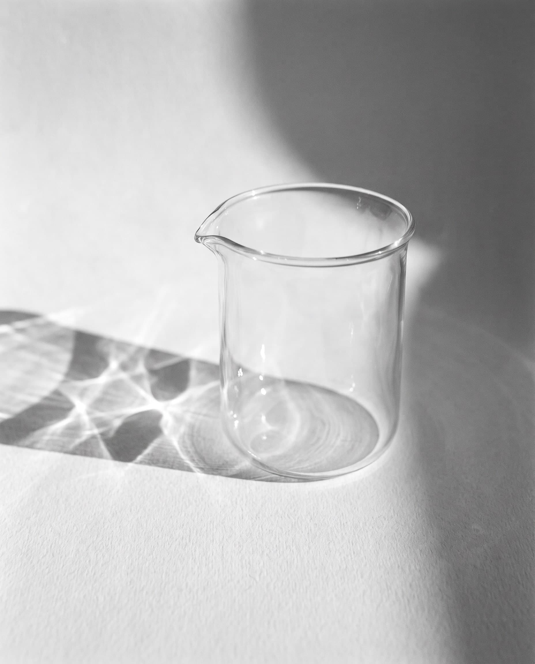 Leeres Becherglas auf heller Fläche, Licht bricht sich im Glas, schwarzweiß